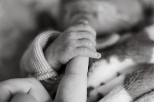 A tender moment is captured in black and white, showing a tiny baby hand gripping an adult finger. The baby is dressed in a knitted garment, suggesting warmth and care. The image conveys a sense of connection and protection.