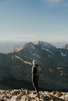 A person wearing outdoor gear, including a hat and backpack, stands on a rocky mountain summit. Rugged mountain peaks stretch into the distance under a clear blue sky, with sunlight casting dramatic shadows on the landscape.