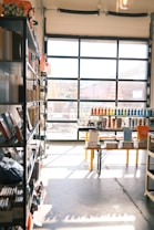 A sunlit store interior with shelves filled with various stationery items. Books and notebooks are neatly arranged on metal shelves to the left. A large glass door at the back allows natural light to illuminate the space, casting shadows on the concrete floor. In the center, a table displays more stationery, including pens organized by color.