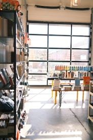 A sunlit store interior with shelves filled with various stationery items. Books and notebooks are neatly arranged on metal shelves to the left. A large glass door at the back allows natural light to illuminate the space, casting shadows on the concrete floor. In the center, a table displays more stationery, including pens organized by color.