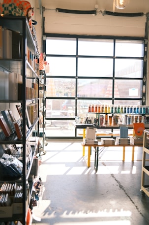 A sunlit store interior with shelves filled with various stationery items. Books and notebooks are neatly arranged on metal shelves to the left. A large glass door at the back allows natural light to illuminate the space, casting shadows on the concrete floor. In the center, a table displays more stationery, including pens organized by color.