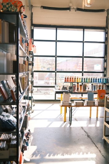 A sunlit store interior with shelves filled with various stationery items. Books and notebooks are neatly arranged on metal shelves to the left. A large glass door at the back allows natural light to illuminate the space, casting shadows on the concrete floor. In the center, a table displays more stationery, including pens organized by color.