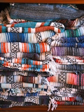 Multiple folded woven blankets stacked neatly on top of one another, featuring various colorful patterns with stripes and geometric designs. The vibrant colors include blues, oranges, reds, and whites, creating a visually striking display.