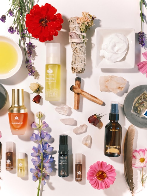 Various wellness and beauty products, including oils in bottles, a bowl of oil, dried flowers, crystals, a smudge stick, and a small dish of cream, are arranged aesthetically on a light surface. The scene is decorated with colorful flowers and herbs.