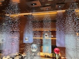 An elegant interior featuring a stunning ceiling installation with hanging crystal-like decorations creating a shimmering effect. Large reflective orbs in silver and red are suspended, enhancing the luxurious ambiance. The lighting is warm, with decorative lights embedded in the ceiling.