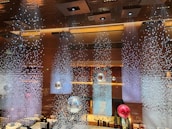 An elegant interior featuring a stunning ceiling installation with hanging crystal-like decorations creating a shimmering effect. Large reflective orbs in silver and red are suspended, enhancing the luxurious ambiance. The lighting is warm, with decorative lights embedded in the ceiling.