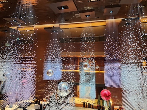 An elegant interior featuring a stunning ceiling installation with hanging crystal-like decorations creating a shimmering effect. Large reflective orbs in silver and red are suspended, enhancing the luxurious ambiance. The lighting is warm, with decorative lights embedded in the ceiling.