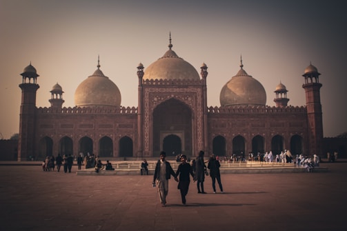 A grand mosque with intricate architectural details and large domes standing prominently. People are walking and gathering in the courtyard, creating a sense of activity. The overall atmosphere is serene and historical.