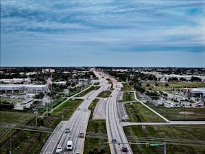 A wide highway stretches into the distance under a cloudy sky, surrounded by green fields and a suburban area with commercial buildings, including car dealerships, and parked vehicles. Traffic is visible on the road with a variety of cars, while American flags are prominently displayed near the businesses.