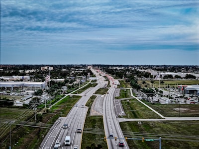 A wide highway stretches into the distance under a cloudy sky, surrounded by green fields and a suburban area with commercial buildings, including car dealerships, and parked vehicles. Traffic is visible on the road with a variety of cars, while American flags are prominently displayed near the businesses.