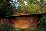 A modernist building with clean, horizontal lines and a flat roof, made primarily of wood and brick. Surrounding the structure are lush green trees and bushes, adding to a serene, natural environment.