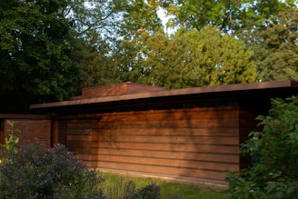 A modernist building with clean, horizontal lines and a flat roof, made primarily of wood and brick. Surrounding the structure are lush green trees and bushes, adding to a serene, natural environment.