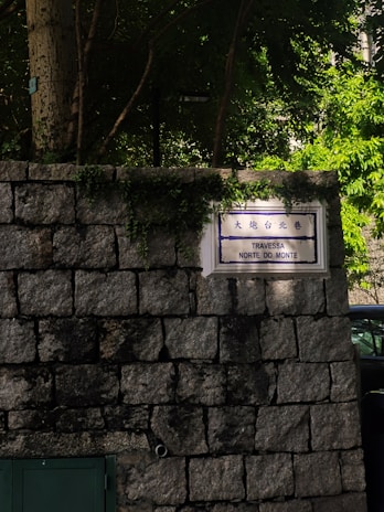 A stone wall with a street sign displaying both Chinese and Portuguese text. The sign is surrounded by green vegetation, and sunlight filters through trees above, casting dappled shadows on the wall. A small section of a dark green door is visible at the bottom left.