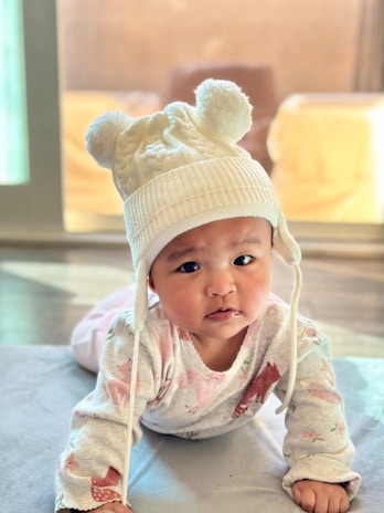 A baby wearing a knitted hat with pom-poms crawls on a soft surface. The child is dressed in a patterned onesie with animal motifs and appears to be indoors, with blurred furniture in the background.