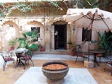 A serene courtyard featuring an old stone wall with arched windows and a large wooden door. There are several potted plants and lush greenery adding to the peaceful atmosphere. Two sets of black wrought iron tables and chairs with red cushions are placed on a light-colored stone floor. A large umbrella provides shade, and a central decorative pot filled with colorful flower petals enhances the overall tranquil and inviting ambiance.