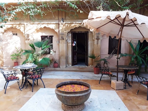 A serene courtyard featuring an old stone wall with arched windows and a large wooden door. There are several potted plants and lush greenery adding to the peaceful atmosphere. Two sets of black wrought iron tables and chairs with red cushions are placed on a light-colored stone floor. A large umbrella provides shade, and a central decorative pot filled with colorful flower petals enhances the overall tranquil and inviting ambiance.