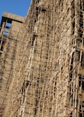 A building under construction is covered with a complex network of bamboo scaffolding. The wooden poles are arranged in a grid-like pattern, providing structural support and access for workers. The sunlight casts shadows on the beige concrete walls, highlighting the texture and lines created by the scaffolding.