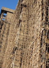 A building under construction is covered with a complex network of bamboo scaffolding. The wooden poles are arranged in a grid-like pattern, providing structural support and access for workers. The sunlight casts shadows on the beige concrete walls, highlighting the texture and lines created by the scaffolding.