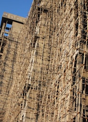 A building under construction is covered with a complex network of bamboo scaffolding. The wooden poles are arranged in a grid-like pattern, providing structural support and access for workers. The sunlight casts shadows on the beige concrete walls, highlighting the texture and lines created by the scaffolding.