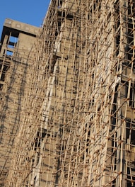 A building under construction is covered with a complex network of bamboo scaffolding. The wooden poles are arranged in a grid-like pattern, providing structural support and access for workers. The sunlight casts shadows on the beige concrete walls, highlighting the texture and lines created by the scaffolding.