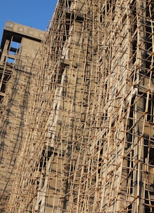 A building under construction is covered with a complex network of bamboo scaffolding. The wooden poles are arranged in a grid-like pattern, providing structural support and access for workers. The sunlight casts shadows on the beige concrete walls, highlighting the texture and lines created by the scaffolding.