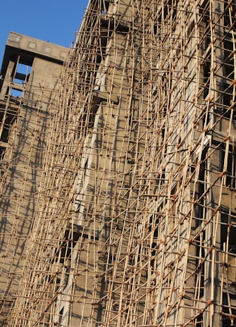 A building under construction is covered with a complex network of bamboo scaffolding. The wooden poles are arranged in a grid-like pattern, providing structural support and access for workers. The sunlight casts shadows on the beige concrete walls, highlighting the texture and lines created by the scaffolding.