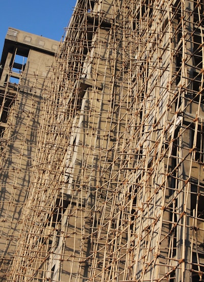 A building under construction is covered with a complex network of bamboo scaffolding. The wooden poles are arranged in a grid-like pattern, providing structural support and access for workers. The sunlight casts shadows on the beige concrete walls, highlighting the texture and lines created by the scaffolding.
