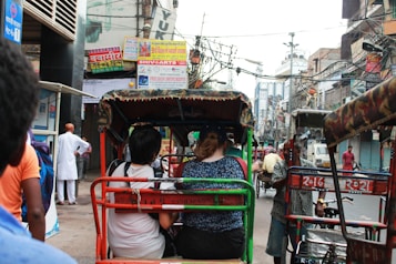 A bustling street scene featuring several rickshaws navigating through a busy urban area. Numerous signs adorn the storefronts and buildings. People, both locals and tourists, are either walking or seated in rickshaws. Overhead, there is a tangle of electrical wires crisscrossing the scene, typical of dense city infrastructure. The atmosphere feels lively and dynamic.