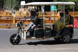 A motorized rickshaw with open sides is traveling on a road, carrying three passengers. The driver is at the front steering, while two passengers sit in the back. Roadside barriers and signs are visible in the background, indicating urban surroundings.