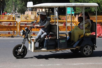 A motorized rickshaw with open sides is traveling on a road, carrying three passengers. The driver is at the front steering, while two passengers sit in the back. Roadside barriers and signs are visible in the background, indicating urban surroundings.