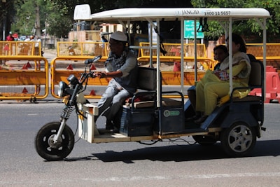 A motorized rickshaw with open sides is traveling on a road, carrying three passengers. The driver is at the front steering, while two passengers sit in the back. Roadside barriers and signs are visible in the background, indicating urban surroundings.