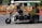 A motorized rickshaw with open sides is traveling on a road, carrying three passengers. The driver is at the front steering, while two passengers sit in the back. Roadside barriers and signs are visible in the background, indicating urban surroundings.