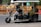A motorized rickshaw with open sides is traveling on a road, carrying three passengers. The driver is at the front steering, while two passengers sit in the back. Roadside barriers and signs are visible in the background, indicating urban surroundings.