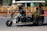 A motorized rickshaw with open sides is traveling on a road, carrying three passengers. The driver is at the front steering, while two passengers sit in the back. Roadside barriers and signs are visible in the background, indicating urban surroundings.