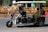 A motorized rickshaw with open sides is traveling on a road, carrying three passengers. The driver is at the front steering, while two passengers sit in the back. Roadside barriers and signs are visible in the background, indicating urban surroundings.