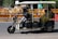 A motorized rickshaw with open sides is traveling on a road, carrying three passengers. The driver is at the front steering, while two passengers sit in the back. Roadside barriers and signs are visible in the background, indicating urban surroundings.