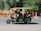 A green motorized rickshaw carrying a group of children dressed in school uniforms, each with colorful backpacks. The rickshaw is moving along a street lined with trees and orange construction barriers.