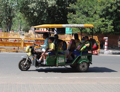 A green motorized rickshaw carrying a group of children dressed in school uniforms, each with colorful backpacks. The rickshaw is moving along a street lined with trees and orange construction barriers.