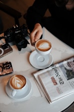A person is holding a cup of coffee with latte art on top. There is another similar cup on the table alongside a camera, a smartphone, and a folded newspaper. The setting suggests a relaxed, informal atmosphere.