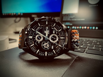A close-up view of a black sports watch with a detailed face, featuring multiple dials and silver markers. The watch is resting on a laptop keyboard, with blurred computer screens in the background. The watch strap is made of braided material with colors including blue, orange, and white.