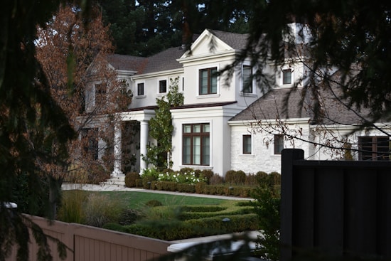 A large, elegant two-story house with a symmetrical design and classical architecture, featuring white walls, a stone facade, and a taupe roof. Surrounded by a well-maintained garden with manicured hedges and a variety of plants. The house is partially obscured by tree branches, suggesting a private, serene environment.