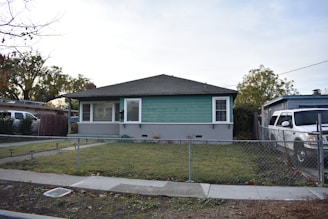 A single-story house with green siding and white trim sits behind a small chain-link fence. The front yard is mostly grass with a narrow concrete path leading to the porch. A white SUV is parked in the driveway beside the house. There are trees in the background and another house visible to the left.