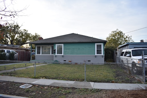 A single-story house with green siding and white trim sits behind a small chain-link fence. The front yard is mostly grass with a narrow concrete path leading to the porch. A white SUV is parked in the driveway beside the house. There are trees in the background and another house visible to the left.
