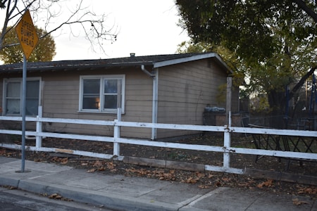 A small, single-story house with beige siding and white-framed windows. A white wooden fence runs along the front yard, with a sidewalk next to it. A 'Speed Bumps Ahead' sign is situated in the foreground. The yard is scattered with fallen leaves and a few large trees are in the background, creating a suburban atmosphere.