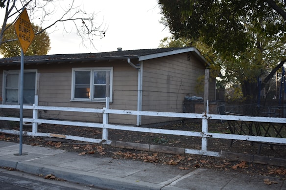 A small, single-story house with beige siding and white-framed windows. A white wooden fence runs along the front yard, with a sidewalk next to it. A 'Speed Bumps Ahead' sign is situated in the foreground. The yard is scattered with fallen leaves and a few large trees are in the background, creating a suburban atmosphere.