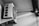 A black and white photograph of an air conditioning unit mounted on a wall. The unit features a large fan and rectangular vents. Shadows and light contrast create a dramatic effect on the wall.