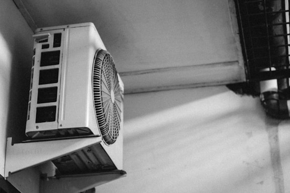 A black and white photograph of an air conditioning unit mounted on a wall. The unit features a large fan and rectangular vents. Shadows and light contrast create a dramatic effect on the wall.