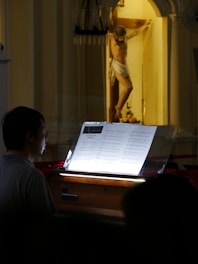 A person is playing or reading sheet music from a music stand with a religious statue of Jesus on the cross in the background, inside a dimly lit church.