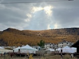 A serene landscape with a foreground of traditional yurts and modern buildings, set against a backdrop of a dense forest on a mountain slope. The sky is overcast with a partial opening that reveals sunlight breaking through.