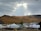 A serene landscape with a foreground of traditional yurts and modern buildings, set against a backdrop of a dense forest on a mountain slope. The sky is overcast with a partial opening that reveals sunlight breaking through.